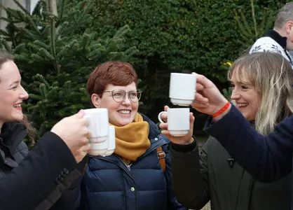 Fünf fröhliche Menschen stoßen vor einem Tannenbaum mit weißen Tassen an, weihnachtliche Stimmung