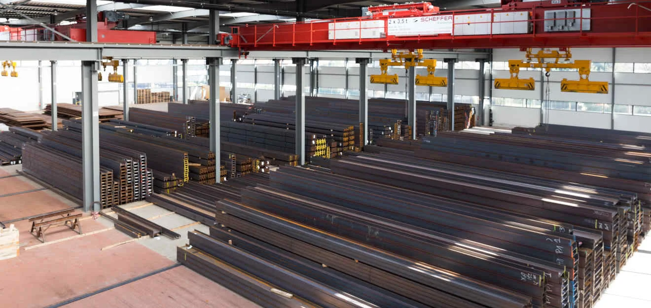 Halle mit gestapelten Stahlprofilen; oben rote Krananlagen mit gelben Greifern und Oberlichtern