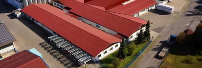 Luftaufnahme von Werkhallen mit roten Dächern; links Solarmodule, rechts Straße mit Lkw