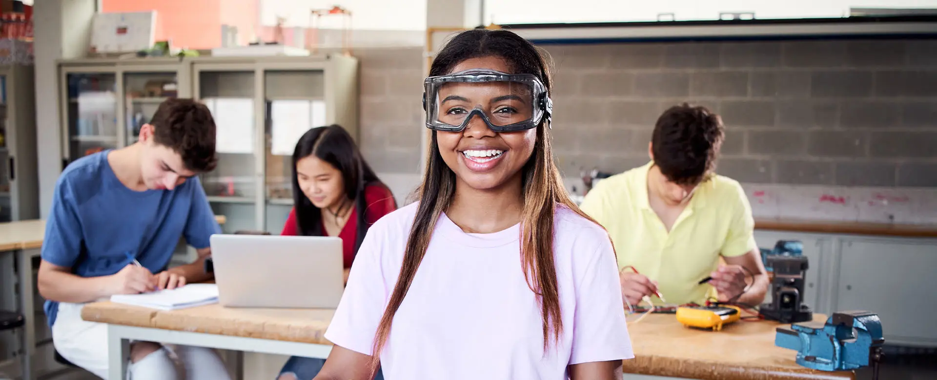 Schülerin mit Schutzbrille im Technikunterricht, lächelnd, Mitschüler im Hintergrund an Projekten