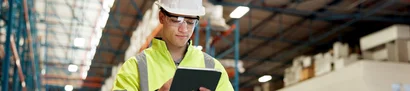 Mitarbeiter mit Helm und gelber Leuchtjacke prüft Logistikdaten auf einem Tablet im Lager.