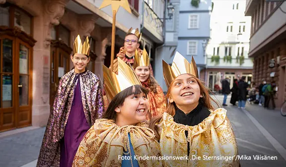 Fünf fröhlich lächelnde Kinder laufen im Sternsinger-Gewand durch eine Altstadt 