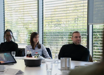 Drei Personen sitzen am Konferenztisch; vor ihnen stehen Flaschen und ein Laptop, hinten Jalousien.