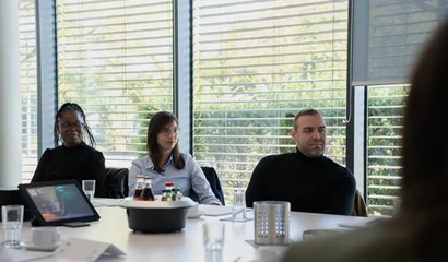 Drei Personen sitzen am Konferenztisch; vor ihnen stehen Flaschen und ein Laptop, hinten Jalousien.