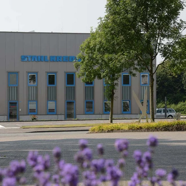Company façade with lettering ‘STAHL KREBS’ Facade with STAHL KREBS lettering, blue window frames; trees in front, parked vehicle on the right.