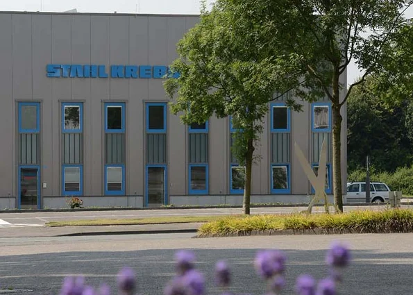 Fassade mit Schriftzug STAHL KREBS, blaue Fensterrahmen; davor Bäume, rechts geparktes Fahrzeug.