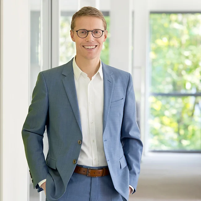 Christoph Hilligen, CEO World Vision Deutschland e. V. Lächelnder Mann mit Brille im blauen Anzug steht in einem hellen Büroflur von World Vision.