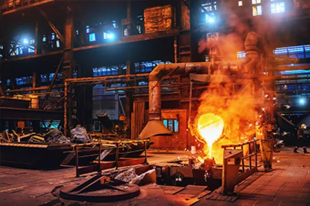 Funken fliegen, zwei Personen stehen in Schutzkleidung neben einem gleißend heißen Schmelztiegel