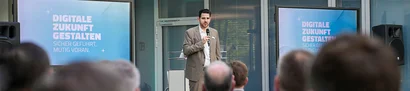 Mann mit Mikrofon vor Publikum; Folie „Digitale Zukunft gestalten“ auf zwei Bildschirmen.