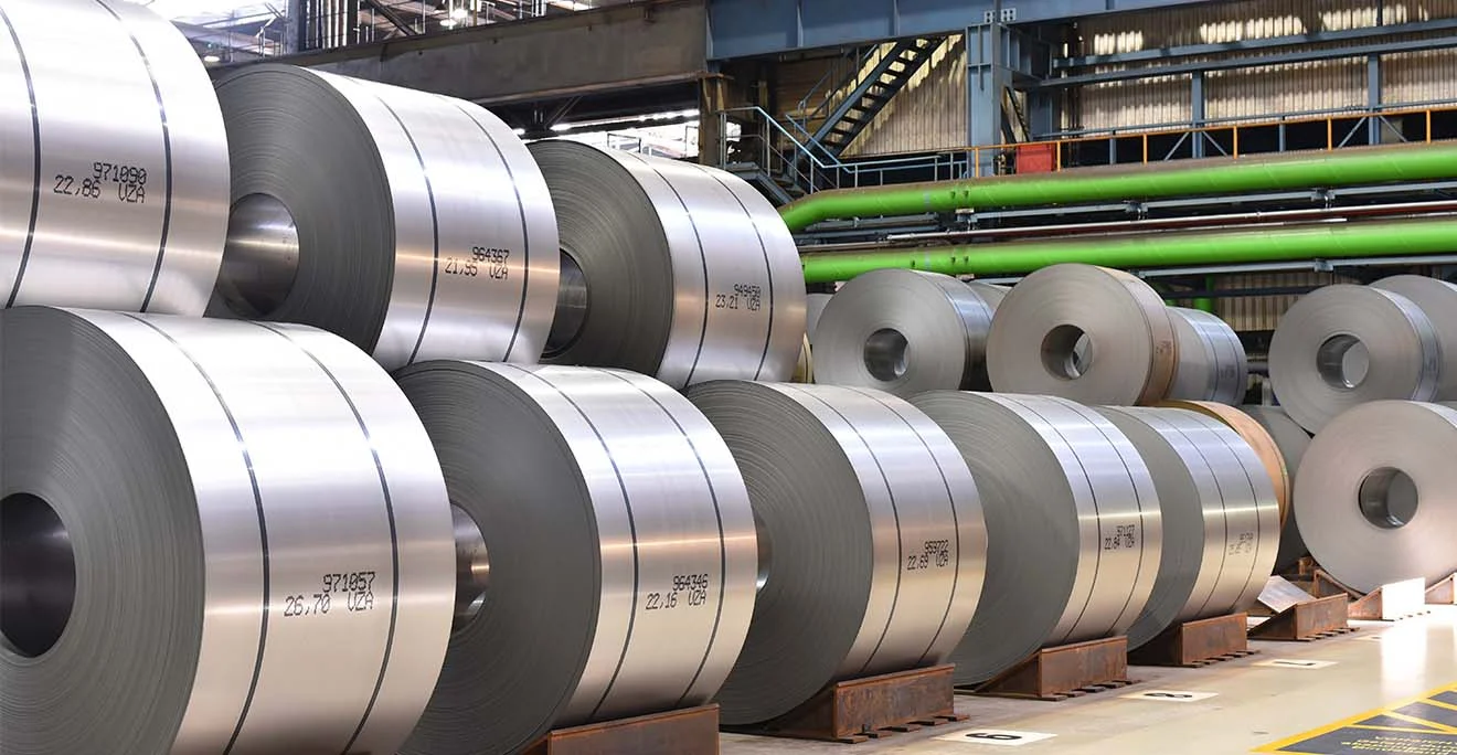 Große Stahl-Coils stehen nebeneinander in einer Werkshalle mit grünen Rohrleitungen im Hintergrund.
