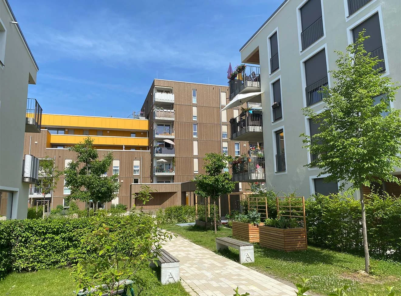 Moderne Wohnanlage mit begrüntem Innenhof, Sitzbänken, Hochbeeten und Balkonen unter blauem Himmel