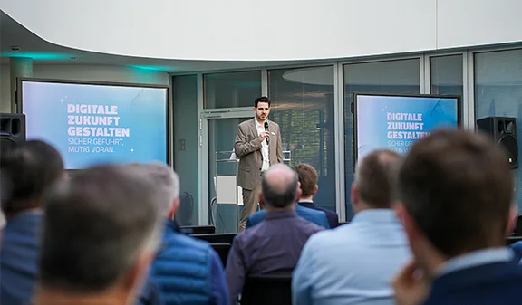 Mann mit Mikrofon vor Publikum; Folie „Digitale Zukunft gestalten“ auf zwei Bildschirmen.