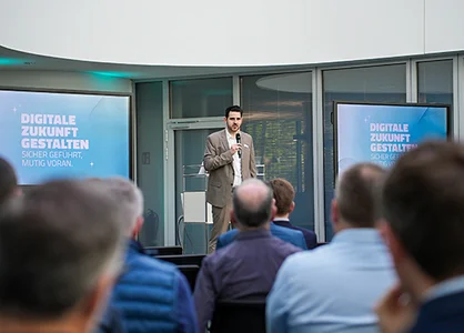 Mann mit Mikrofon vor Publikum; Folie „Digitale Zukunft gestalten“ auf zwei Bildschirmen.