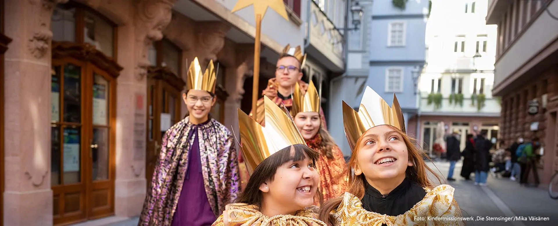 Fünf fröhlich lächelnde Kinder laufen im Sternsinger-Gewand durch eine Altstadt 