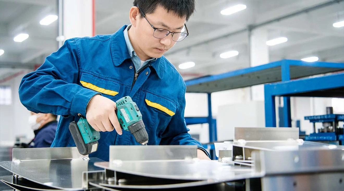 Employee screws metal components together with a cordless screwdriver at an assembly station.