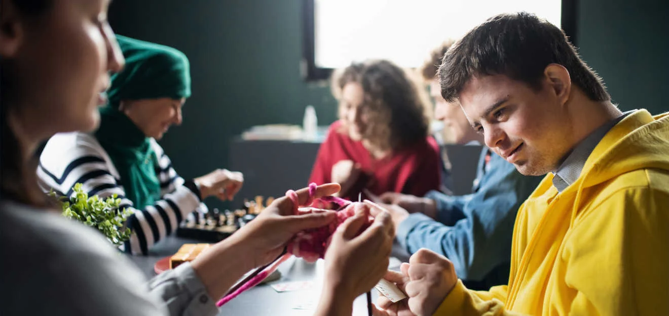 Gemeinsame Freizeitaktivität in der Evangelischen Stiftung Neuerkerode Junger Mann mit Down-Syndrom sitzt mit Gruppe beim Spielen und Handarbeiten an einem Tisch.