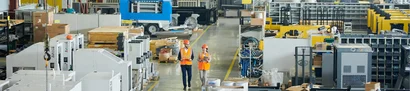 Panoramic view through a production hall with machines, shelves and materials; two people.