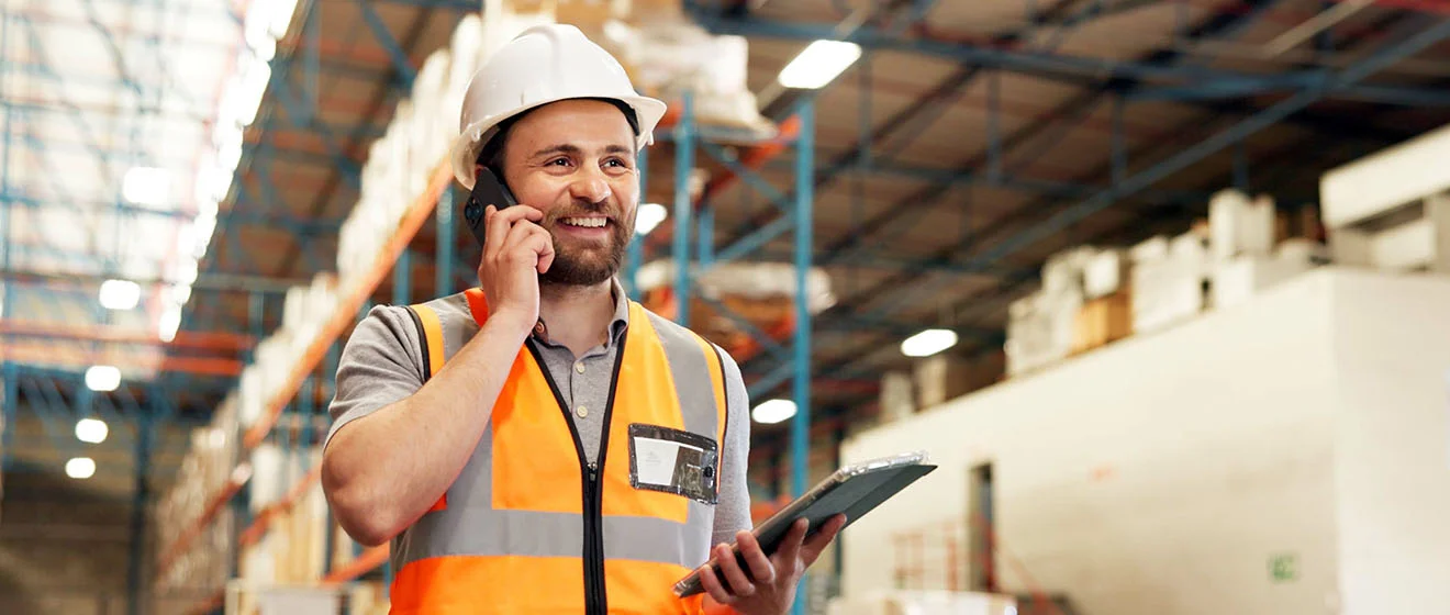 Wie digital ist Ihre Logistik aufgestellt? Lagerlogistik-Mitarbeiter mit Helm und Warnweste telefoniert und hält Tablet im Hochregallager.