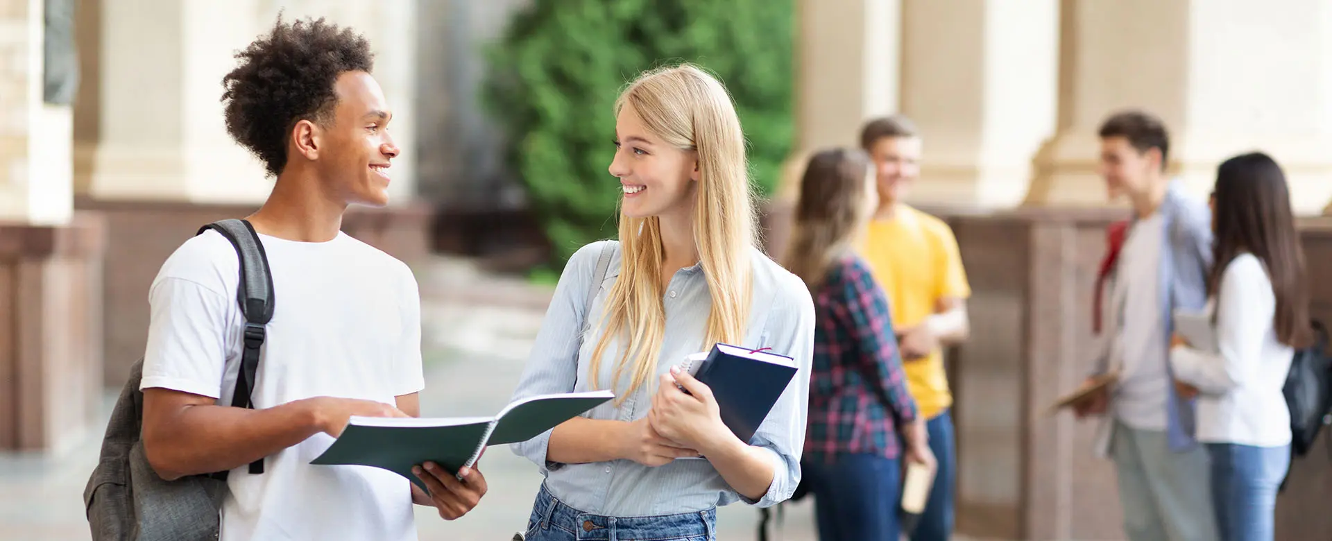 Zwei lächelnde Studierende mit Heften in den Händen unterhalten sich vor einer belebten Campus-Szene.