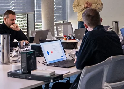 Start der neuen Trainee-Gruppe: Onboarding-Runde Archivfoto: Gruppe mit Laptops am Tisch; IT-Mitarbeiter steht vorn neben Bildschirm und Flipchart.