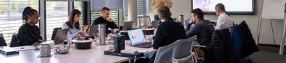 Start der neuen Trainee-Gruppe: Onboarding-Runde Archivfoto: Gruppe mit Laptops am Tisch; IT-Mitarbeiter steht vorn neben Bildschirm und Flipchart.