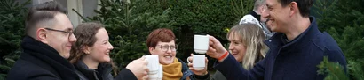 GOB-Weihnachtsevent 2024 auf dem Mengelshof (Kempen) | 1905 x 425 Pixel Fünf fröhliche Menschen stoßen vor einem Tannenbaum mit weißen Tassen an, weihnachtliche Stimmung