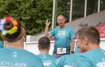 Ein Mann im GOB-Laufshirt spricht vor dem Team, im Hintergrund die Tribüne der Galopprennbahn