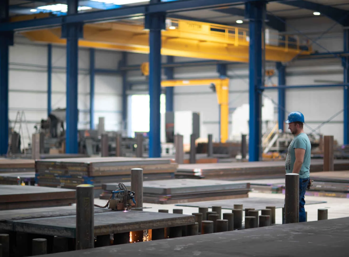 Halle mit gelben Kranbahnen; Arbeiter mit blauem Helm beobachtet Schneidanlage mit Funken.