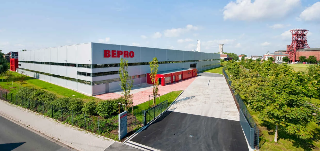 BEPRO-Unternehmenssitz – Außenansicht mit Logo Graues Firmengebäude mit rotem BEPRO-Schriftzug; vorn Weg und Bäume, rechts Industrieanlage