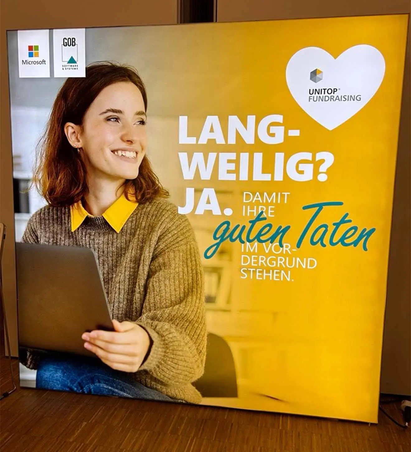 Plakat auf GOB-Stand auf dem DFK 2024; lächelnde Frau am Laptop vor fröhlichem gelben Hintergrund mit unitop-Fundraising-Logo von einem Herzen umrandet am oberen rechten Rand