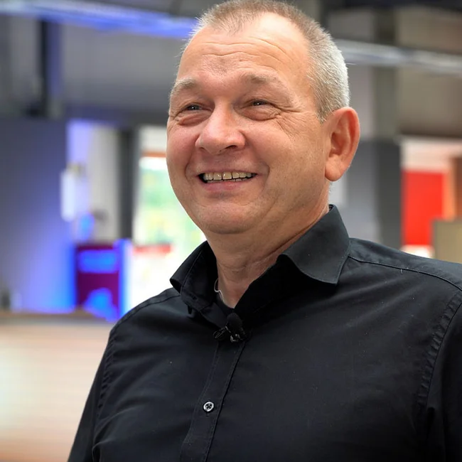 Michael Kusenberg – CIO & Prokurist der Teba GmbH Porträt eines lächelnden Mannes im schwarzen Hemd vor moderner Büroumgebung, halbnah.