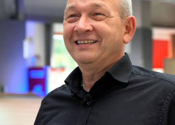 Portrait of a smiling man in a black shirt in front of a modern office environment, medium close-up.