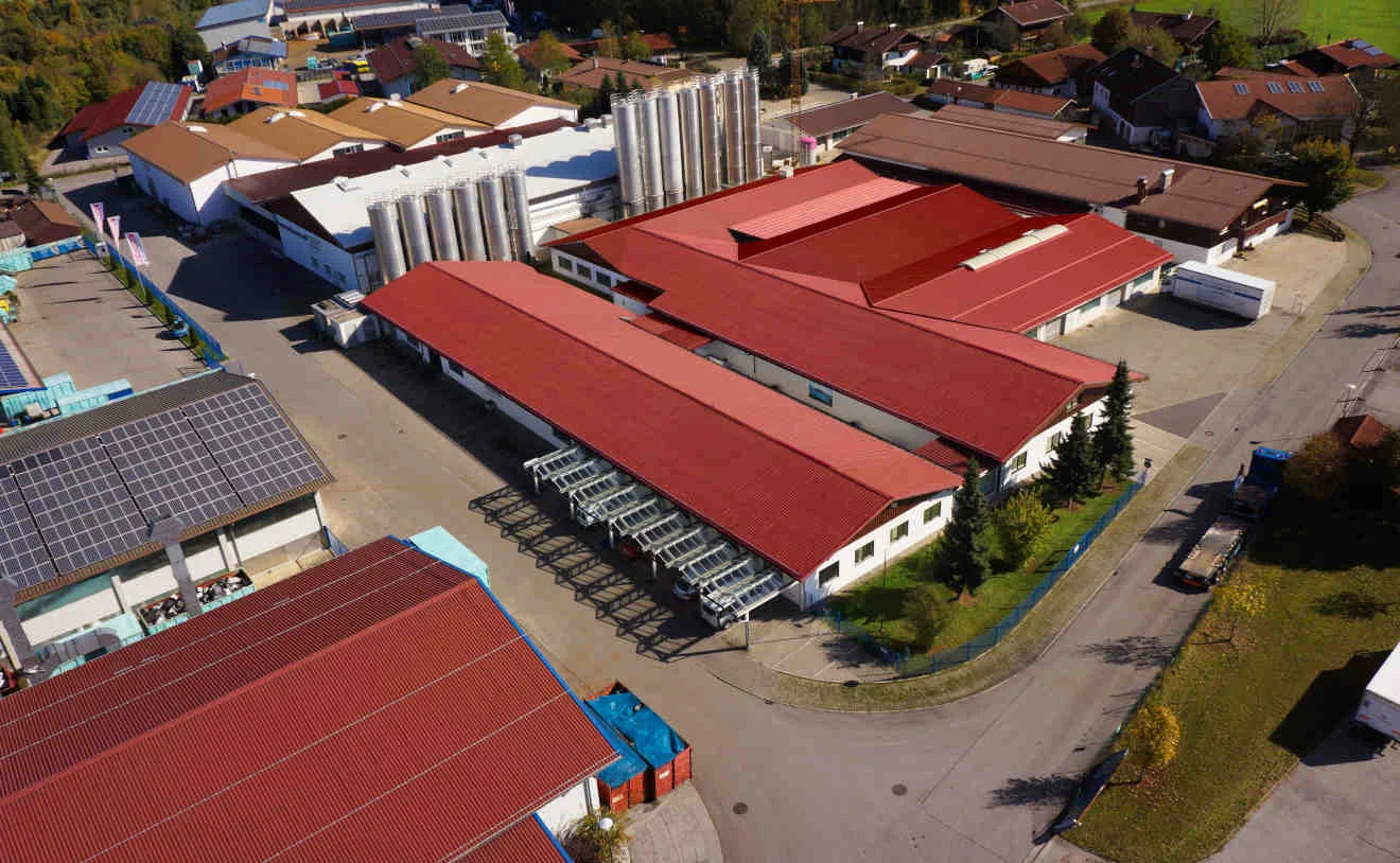 Oldenburg Kunststoff-Technik – Standort Chieming (Werk I) Luftaufnahme von Werkhallen mit roten Dächern; links Solarmodule, rechts Straße mit Lkw