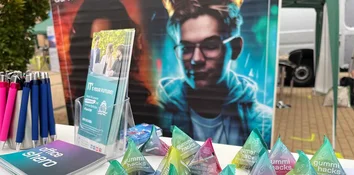 Stifte, Prospekt „IT’S YOUR FUTURE!“ und pyramidenförmige Gummibärchenbeutel vor Posterwand