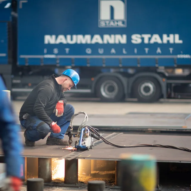 Arbeiter kniet mit Brennschneider auf Stahlplatte; Funken; Lkw mit NAUMANN STAHL IST QUALITÄT.
