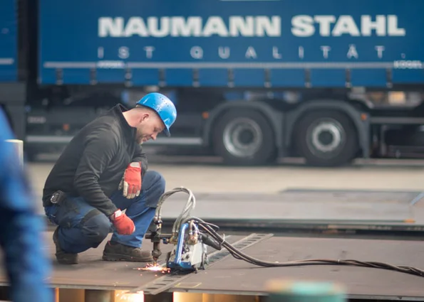 Worker with torch kneeling on steel plate; sparks; lorry with inscription NAUMANN STAHL IST QUALITÄT
