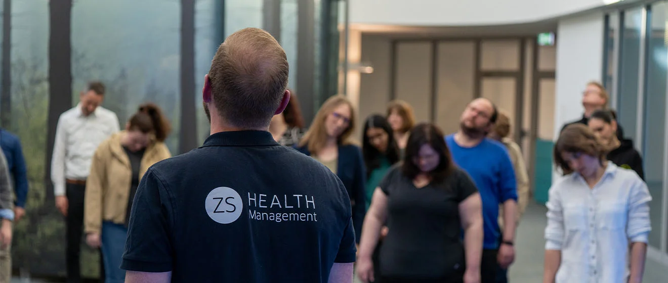 Gruppe macht eine Dehnübung; ein Trainer steht vorne und leitet die Bewegung an.