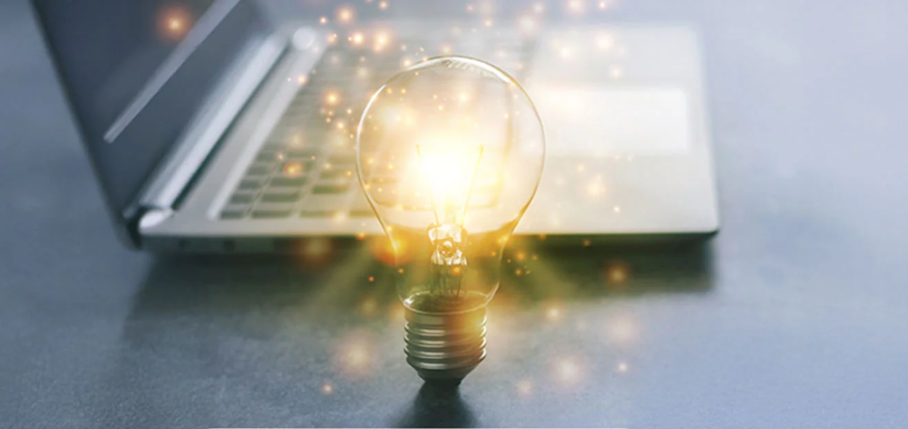 Symbolbild: Leuchtende Glühbirne mit Lichtpartikeln vor einem Laptop auf Tisch.