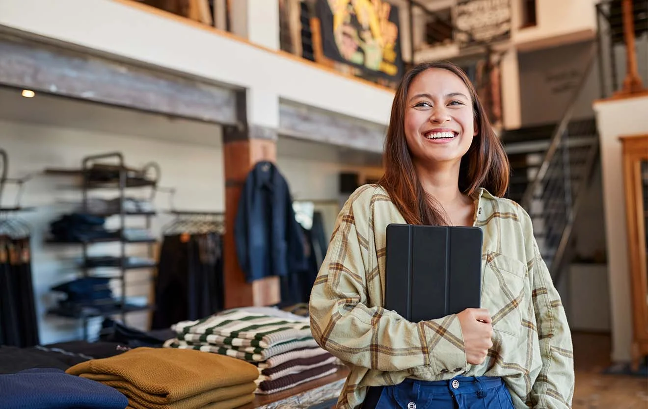 Digitale Unterstützung im Modegeschäft mit ERP-Lösung Lachende Frau steht mit Tablet in einem Modegeschäft vor gefalteter Ware.