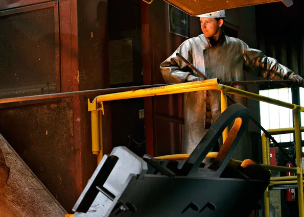 Foundry worker in protective coat at railing; liquid metal glows from a ladle below