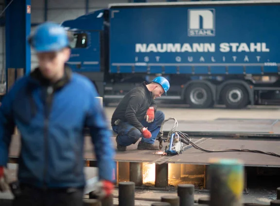 Mitarbeiter kniet an Brennschneidgerät; Funken leuchten; hinten Lkw mit NAUMANN STAHL.