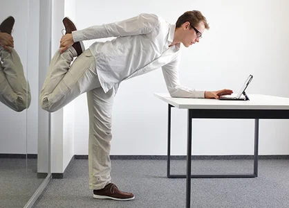 Gesundheit am Arbeitsplatz: Mann macht Dehnübung am Schreibtisch, Laptop daneben