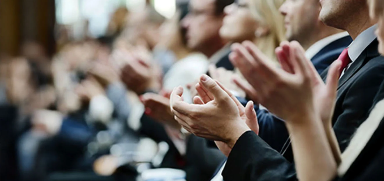 Symbolbild: Hände klatschen in einem Publikum, Menschen im Anzug unscharf im Hintergrund.