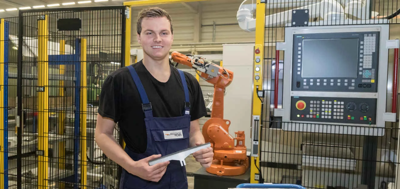 Mitarbeiter in der Produktion von Winterhoff Picard mit Maschinenmesser und Industrieroboter Mitarbeiter von Winterhoff Picard hält Maschinenmesser vor Industrieroboter und Steuerung.