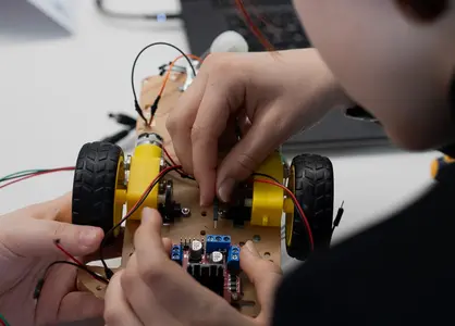 Eine Schülerin und ein Schüler arbeiten an einem kleinen Roboter-Fahrzeug und schließen dünne Kabel an. Auf dem Tisch liegt ein Laptop.