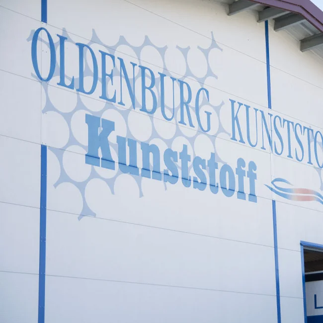 Produktionswerk 1 der Oldenburg Kunststoff-Technik GmbH in Chieming/Bayern Hallenfassade mit blauem Schriftzug „OLDENBURG KUNSTSTOFF“ und grafischen Kreisflächen.