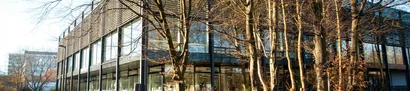 Die Bluebox der Hochschule Bochum bei Tageslicht | 1905 x 425 Pixel Modernes Gebäude mit großen Glasfassaden, umgeben von kahlen Bäumen an einem sonnigen Herbsttag.