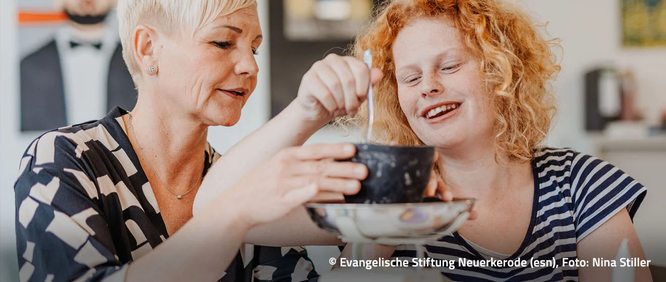 Alltag mit Unterstützung – Copyright: Evangelische Stiftung Neuerkerode, Foto: Nina Stiller Eine Frau unterstützt ein lachendes Mädchen beim Essen am Tisch.