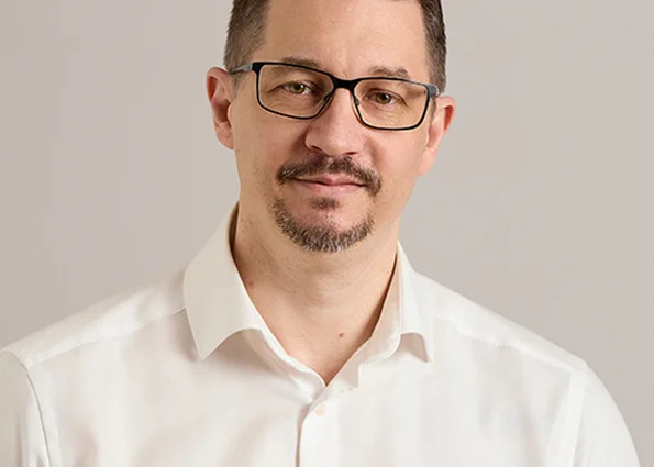 Porträt eines dunkelhaarigen Mannes mit Brille im weißen Hemd vor neutralem Hintergrund.