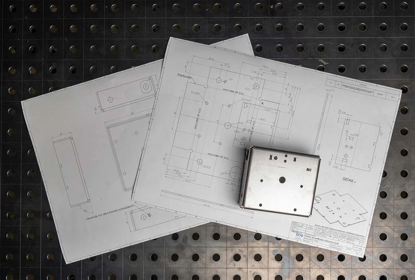 Technische Zeichnungen mit Präzisionsmaßen & ein passendes Metallbauteil auf gelochter Arbeitsfläche